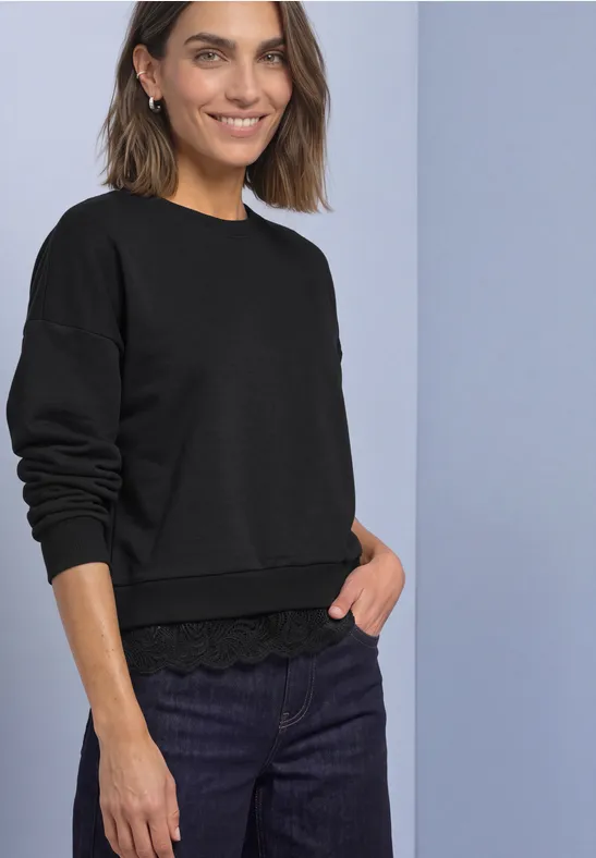 Frau mit kurzem braunem Haar trägt schwarzen Pullover mit Spitzenbund und dunklen Jeans, sie lächelt.