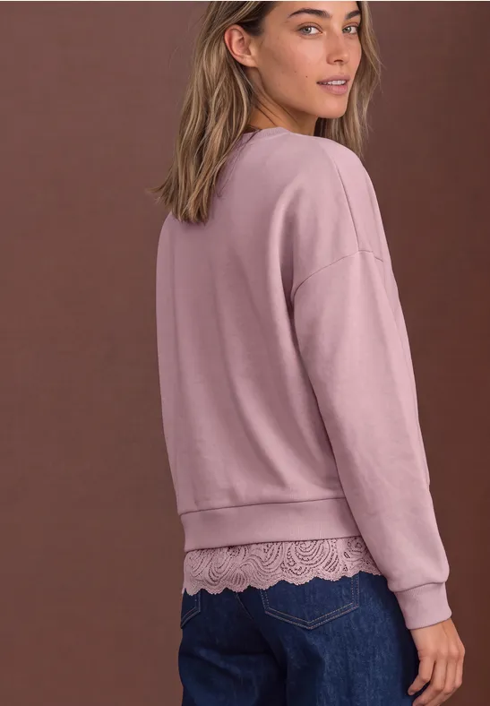 Frau von hinten in rosa Sweatshirt mit Spitzenabschluss über blauen Jeans vor braunem Hintergrund.