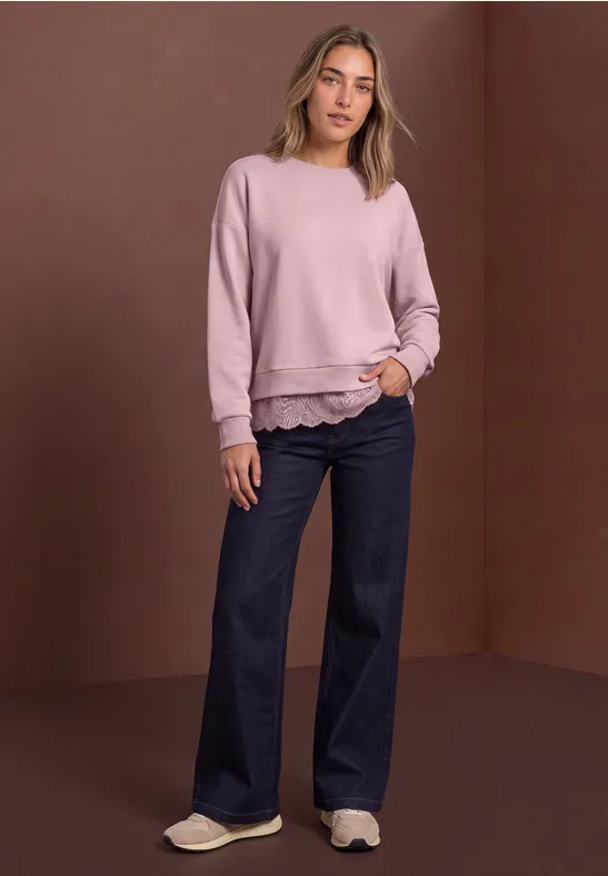 Frau in rosa Sweatshirt und weiten Jeans steht vor braunem Hintergrund, lässige Pose mit Händen in Taschen.