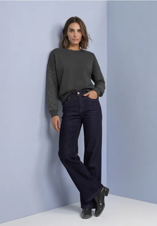 Modell mit dunkelgrünem Pullover, weiten Jeans und schwarzen Boots vor schlichtem hellblauem Hintergrund.