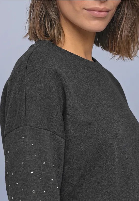 Halbporträt einer Frau in dunkelgrauem Pullover mit kleinen Metallnieten am Ärmel.