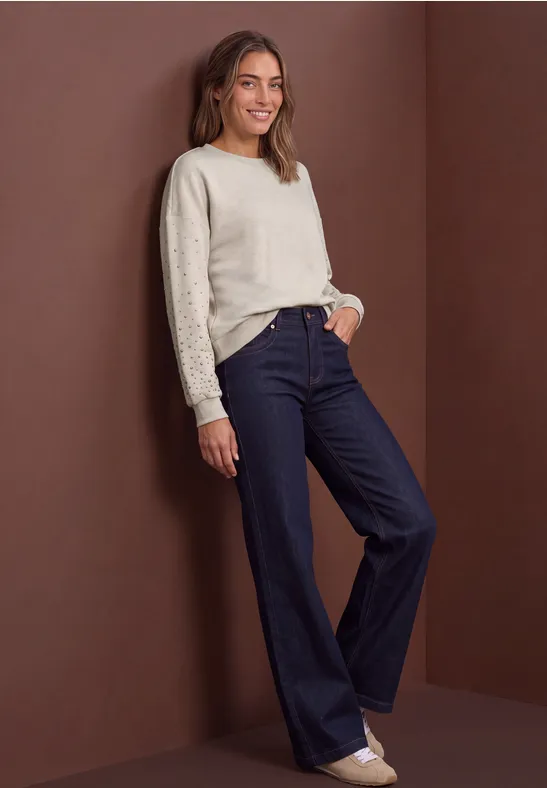 Femme en pull crème et jean évasé foncé appuyée de façon décontractée contre un mur.