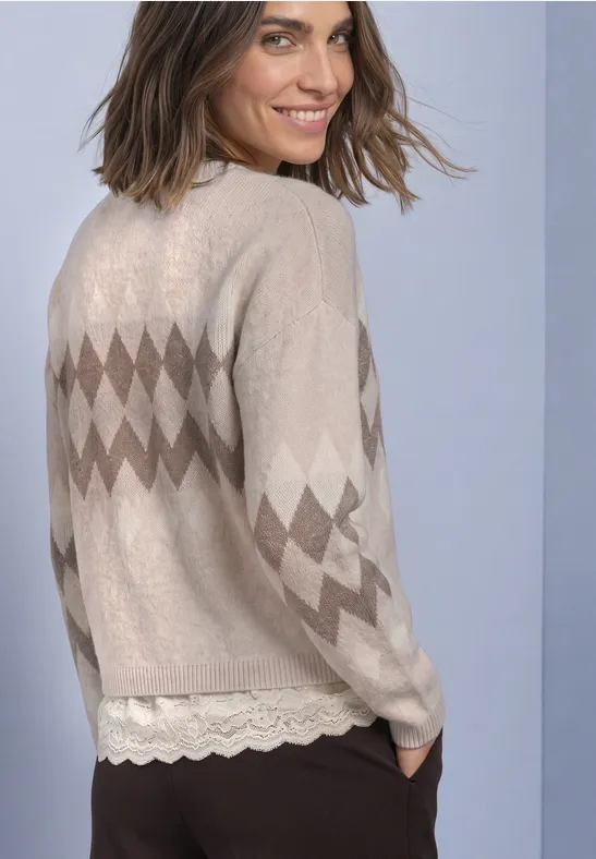 Dame in beiger Strickjacke mit Rautenmuster lächelt über die Schulter, darunter Spitzenbluse.