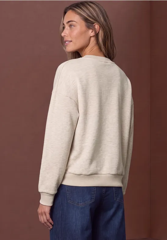 Eine Frau von hinten in beigem Pullover und blauen Jeans vor braunem Hintergrund.