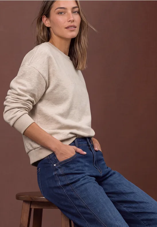 Eine Frau in beigem Pullover und blauen Jeans lehnt lässig auf einem Hocker vor braunem Hintergrund.