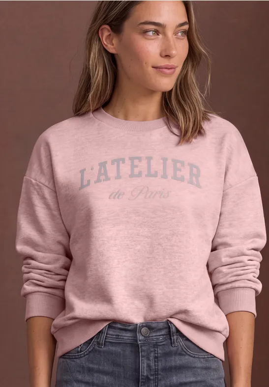 Eine Frau trägt ein rosafarbenes Sweatshirt mit Aufdruck 'L'ATELIER de Paris' kombiniert mit Jeans.