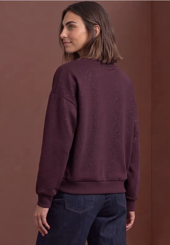 Frau mit mittellangen braunen Haaren in lila Sweatshirt und dunklen Jeans, seitlich von hinten aufgenommen.