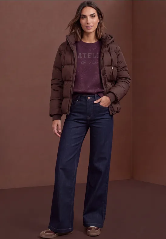 Eine Frau steht vor braunem Hintergrund und trägt eine dunkelbraune Daunenjacke, weite Jeans und Pullover.