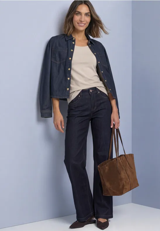 Frau trägt Jeansjacke, beiges Top, weite dunkle Jeans, schwarze Schuhe und braune Tasche vor hellem Hintergrund.