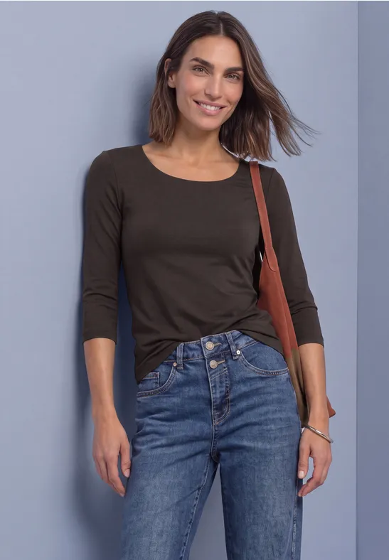 Eine Frau in schwarzem 3/4-Ärmel-Shirt und blauen Jeans mit Tasche, lächelnd vor grauem Hintergrund.
