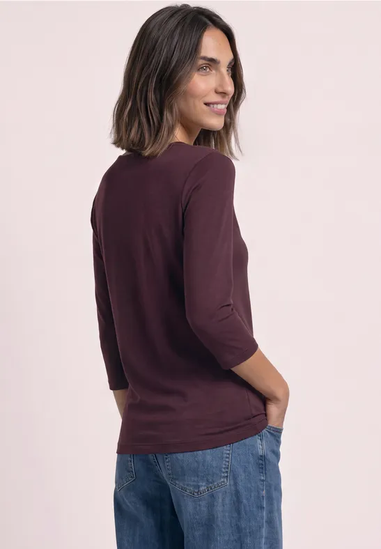 Femme en t-shirt bordeaux 3/4 manches et jean, souriante, regard par-dessus l’épaule, mains dans les poches.