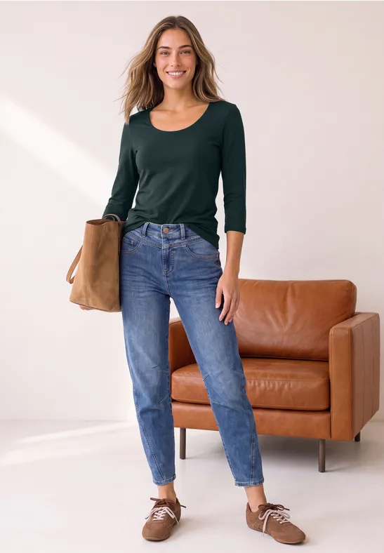 Jeune femme souriante dans un salon lumineux, portant un top vert, un jean bleu, des chaussures et tenant un sac.