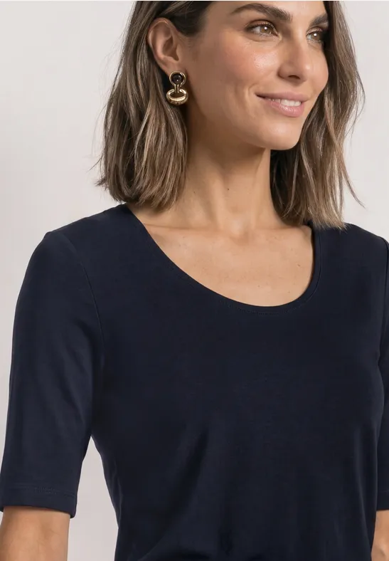 Femme aux cheveux mi-longs portant un t-shirt noir simple à encolure ronde et manches mi-longues, boucles d’oreilles dorées.