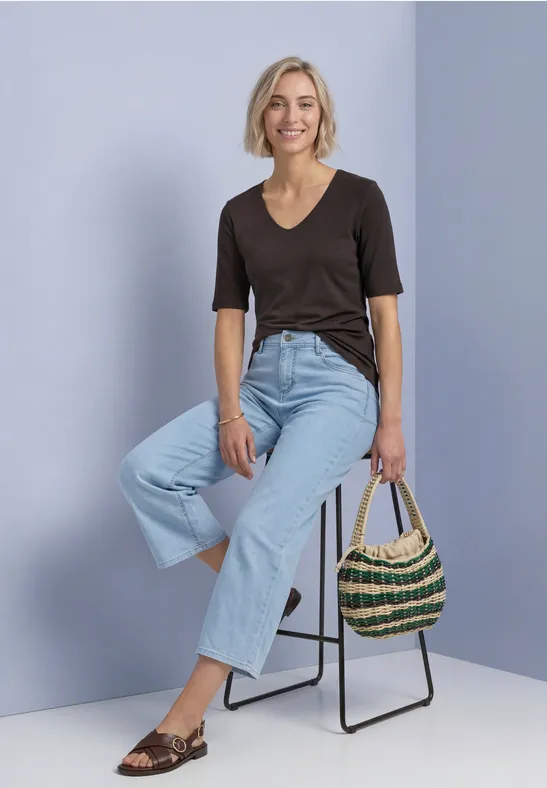 Frau in schwarzem T-Shirt und hellblauen Jeans sitzt auf Hocker, hält eine geflochtene Tasche, lächelnd.