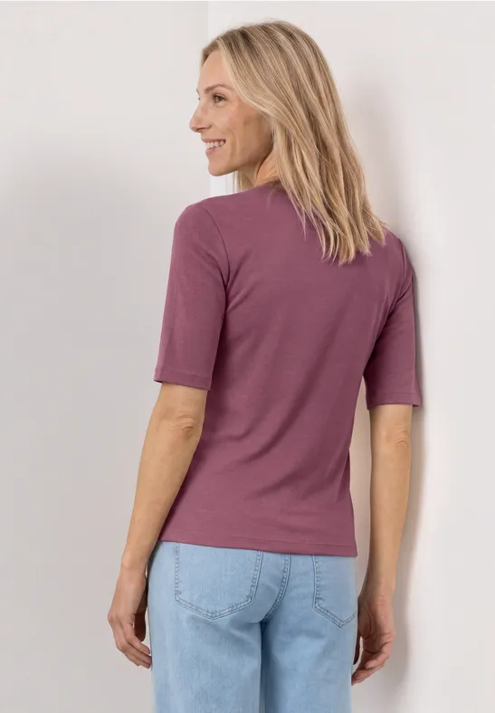 Frau von hinten in violettem T-Shirt und hellblauen Jeans vor neutralem Hintergrund.