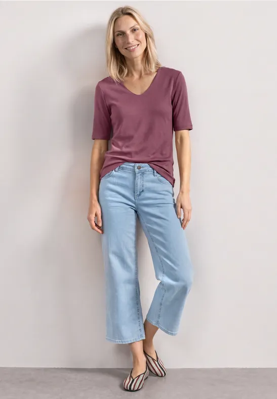 Frau lehnt in lila T-Shirt, hellblauen Culotte-Jeans und gestreiften Sandalen an weißer Wand