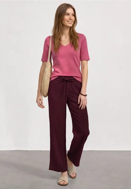 Vrouw in roze T-shirt, losse wijnrode broek, sandalen en strooien tas voor witte achtergrond.