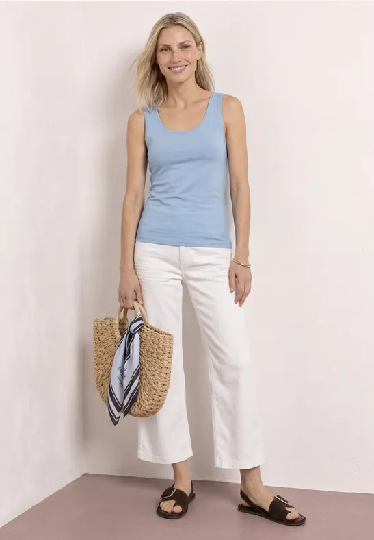 Vrouw met lichtblauw hemdje, witte culotte, zwarte sandalen en rieten tas met sjaal, witte achtergrond.