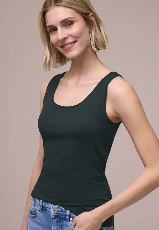 Een persoon met kort blond haar draagt een zwart mouwloos topje en een blauwe spijkerbroek.