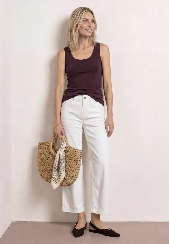 Vrouw in paars top, witte wijde cropped broek, bruine puntschoenen en rieten tas.
