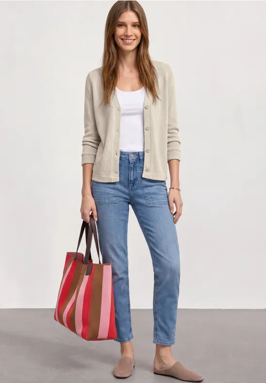 Vrouw in licht vest, wit topje, blauwe jeans, beige schoenen en kleurrijke gestreepte tas.