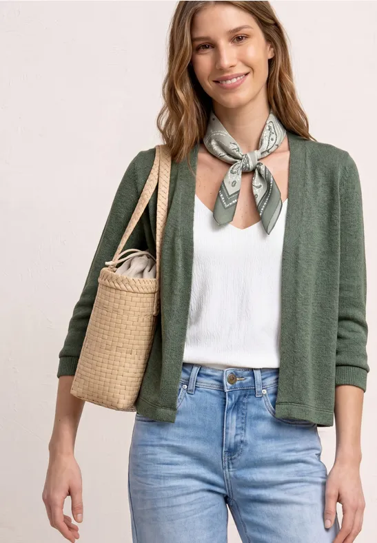 Femme en gilet vert, top blanc et jean, foulard au cou et sac en osier.