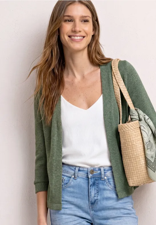 Femme en gilet vert, top blanc et jean, portant un sac en paille tressée à l'épaule