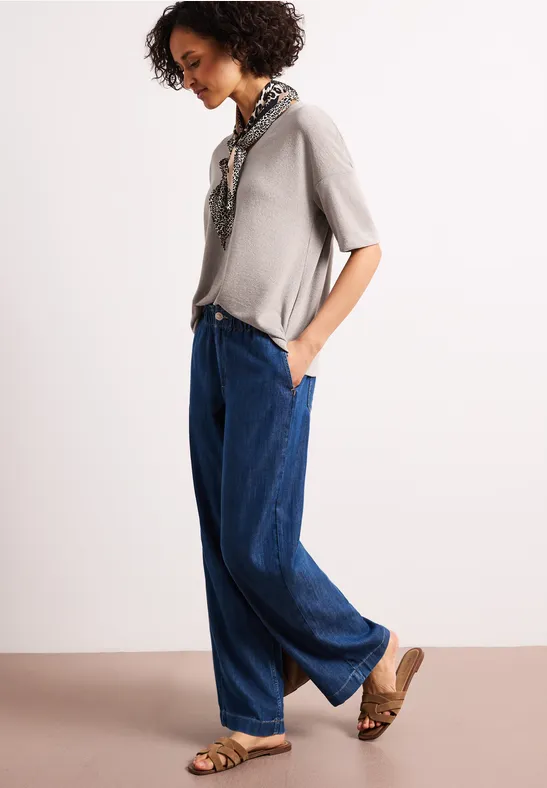 Vrouw zijaanzicht met grijs shirt, wijde blauwe jeans, leren sandalen en zijden sjaal.