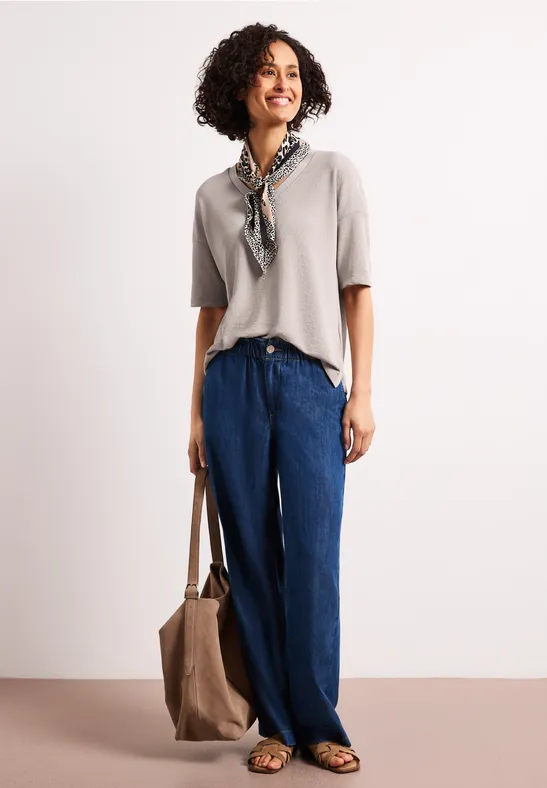 Vrouw voor witte achtergrond, draagt beige top, wijde blauwe broek, sjaal en tas