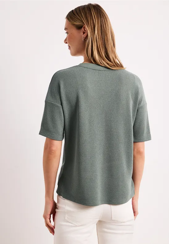 Vrouw van achteren met groen T-shirt met korte mouwen en witte broek tegen een witte achtergrond.