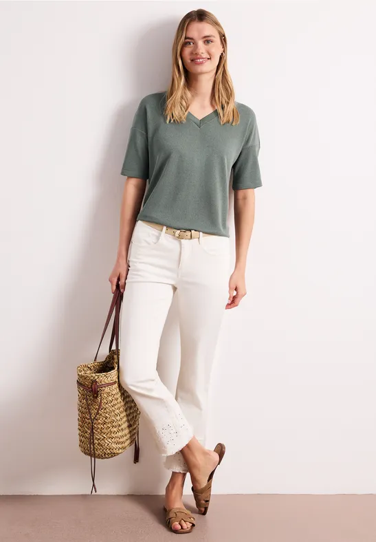 Vrouw met groen T-shirt, witte broek, bruine sandalen, draagt een rieten tas tegen een neutrale achtergrond.