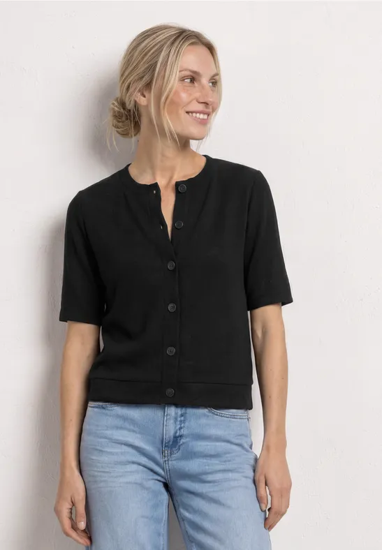 Eine Frau trägt eine schwarze Kurzarm-Strickjacke mit Knöpfen und blaue Jeans, vor neutralem Hintergrund.