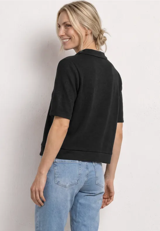 Frau steht seitlich, trägt schwarzen Kurzarm-Pullover und helle Jeans, Blick vom Betrachter abgewandt.