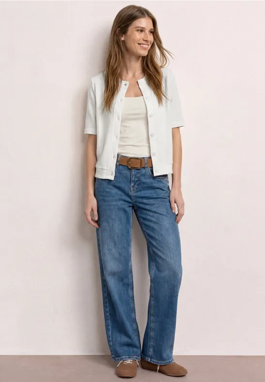 Frau in weißer Kurzjacke, weißem Top, blauen Jeans und braunen Slippern.