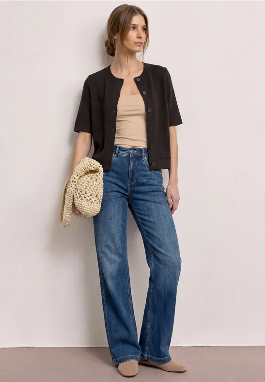 Femme portant un top beige, une veste noire courte, un jean large bleu et un sac tressé.