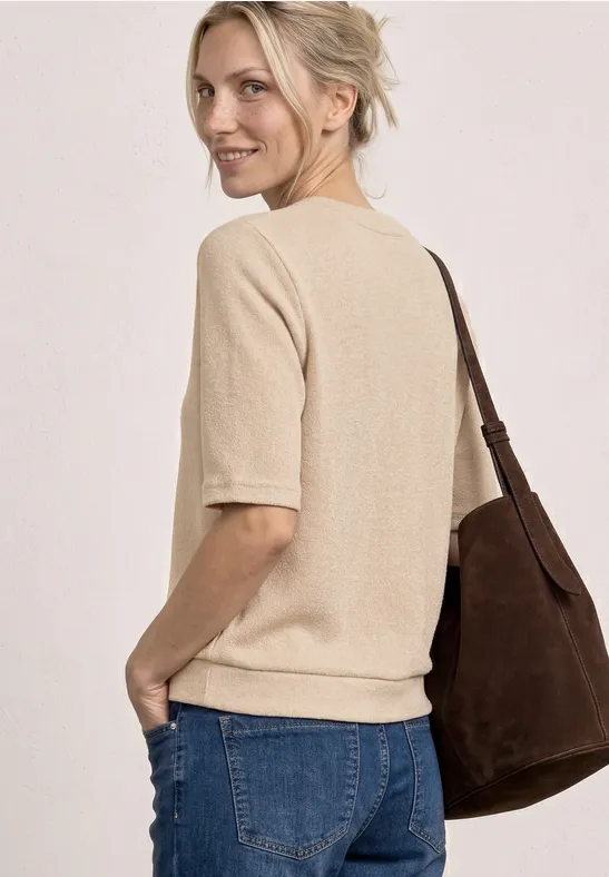 Vrouw in een beige trui met korte mouwen, blauwe jeans en een bruin schoudertasje, zijwaarts pose.