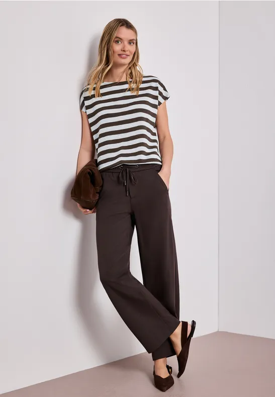 Femme appuyée contre un mur, t-shirt rayé, pantalon ample noir, sandales et pochette en main.