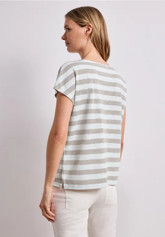 Vrouw van opzij met de rug naar de camera, draagt een grijs-wit gestreept T-shirt en een lichte broek.