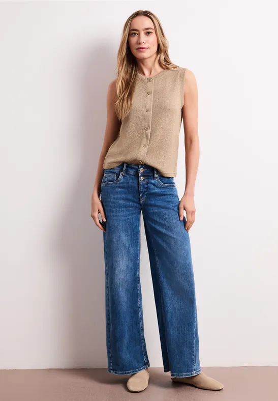 Vrouw in beige top met knopen, wijde blauwe jeans en lichte sloffen voor een witte muur.