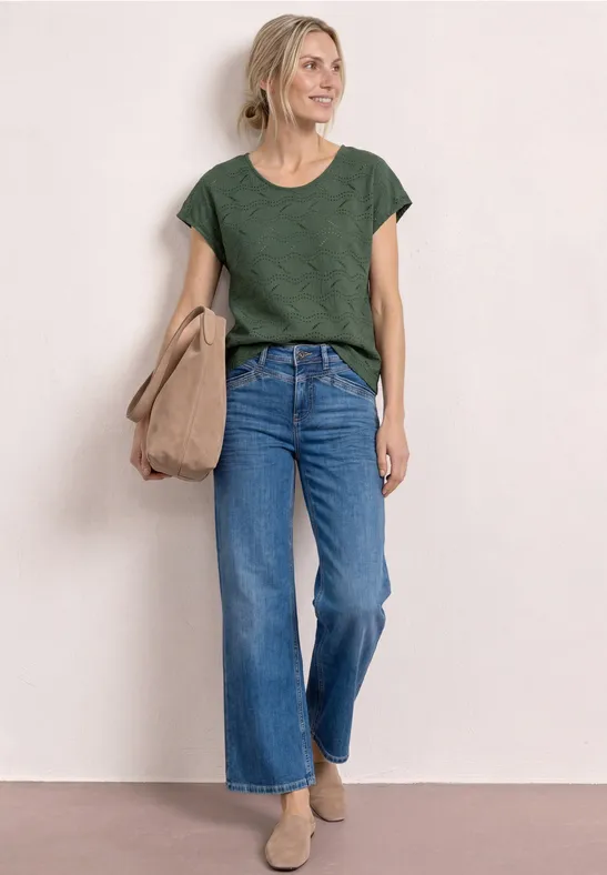 Vrouw met groen t-shirt, brede blauwe jeans en beige tas tegen een effen achtergrond.
