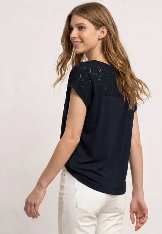 Vrouw van achteren in zwarte kanten top en witte broek tegen lichte achtergrond.