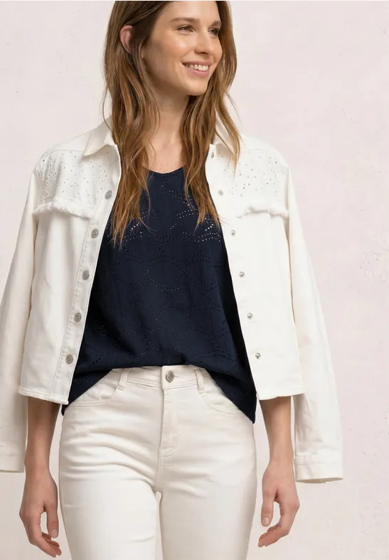 Vrouw in witte spijkerjas, witte broek en donker topje voor een neutrale achtergrond.