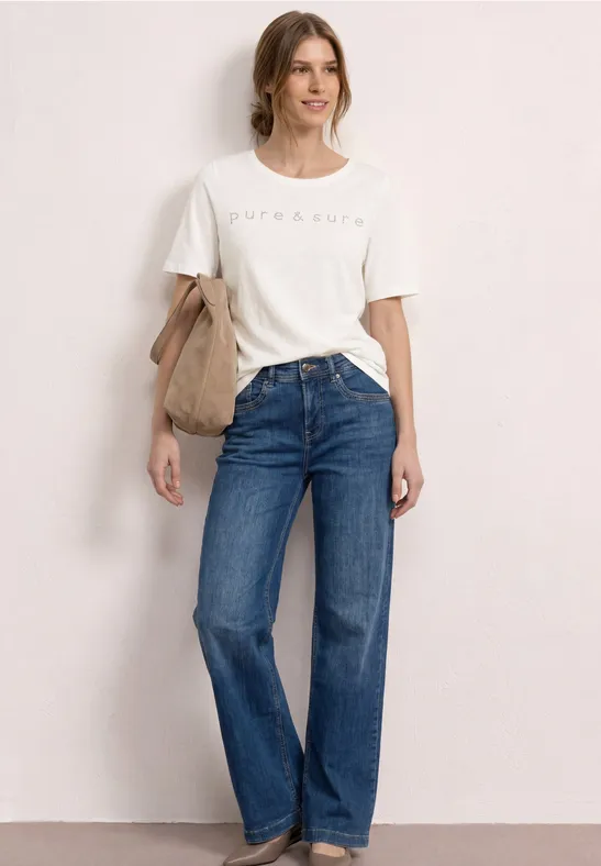 Vrouw voor neutrale achtergrond met wit T-shirt, blauwe jeans en beige schoudertas.
