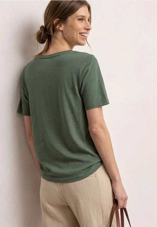 Frau von hinten in olivgrünem T-Shirt und beige Hose, mit kleiner Tasche in der Hand.