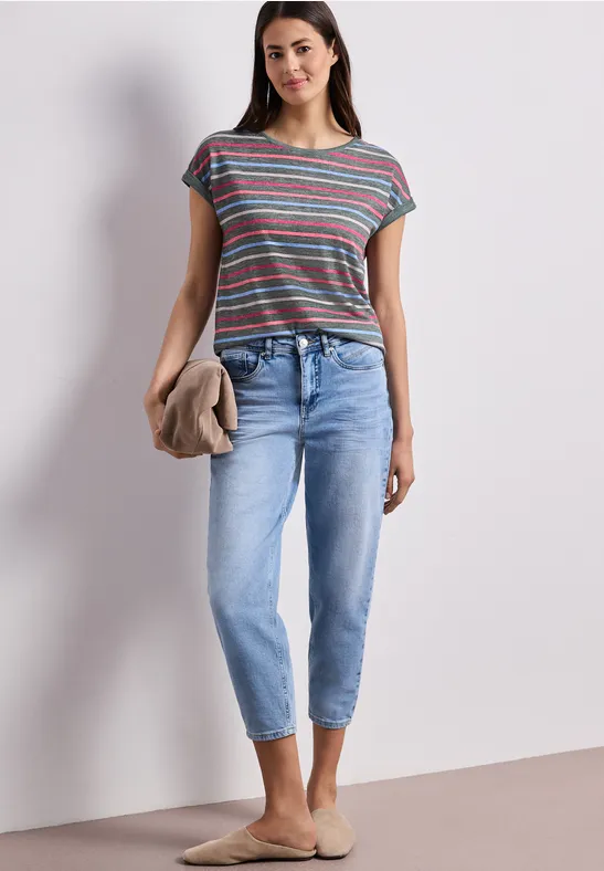 Vrouw in gestreept T-shirt, lichte jeans, houdt een trui, draagt beige instappers voor neutrale achtergrond.