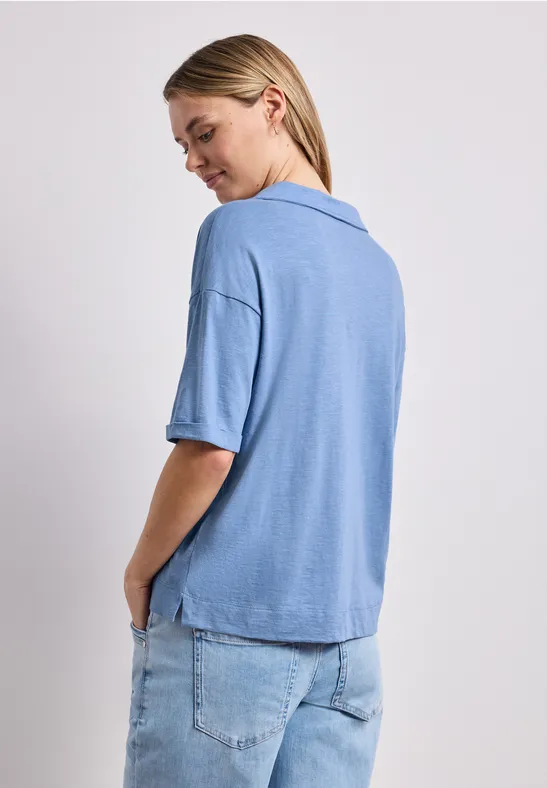 Vrouw van achteren in lichtblauw kort mouw poloshirt en spijkerbroek tegen witte achtergrond.