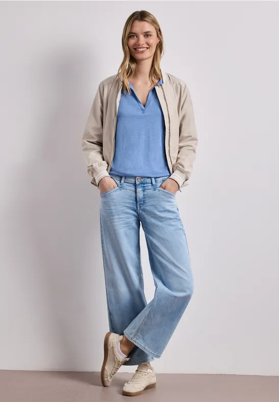 Persoon in beige jack, blauw shirt, wijde spijkerbroek en sneakers, ontspannen met handen in de zakken.