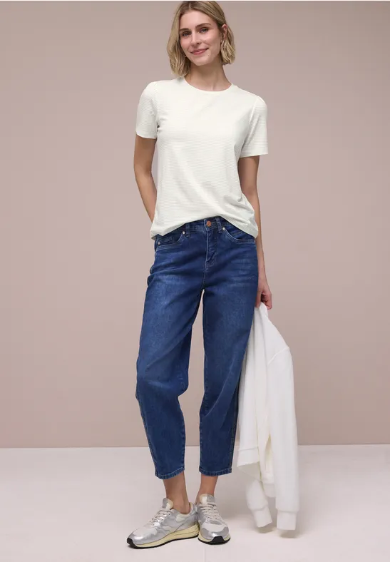 Persoon in wit T-shirt, blauwe high-waist jeans en sneakers, houdt witte jas vast voor neutrale achtergrond.