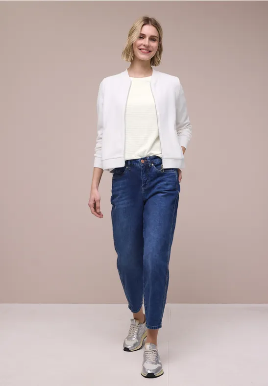 Een vrouw draagt een witte jas, licht T-shirt, blauwe jeans en zilveren schoenen voor een beige achtergrond.