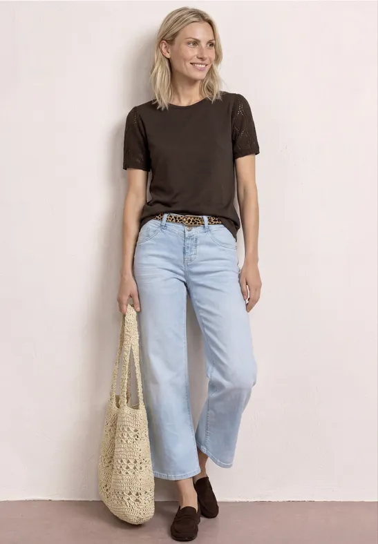 Vrouw in zwart T-shirt, wijde lichtblauwe jeans, bruine riem en gehaakte tas voor een lichte muur.